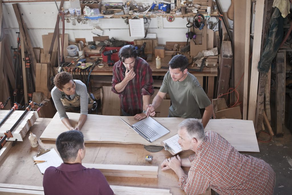 A team of multi-racial factory workers problem solving at a work station in a woodworking factory.